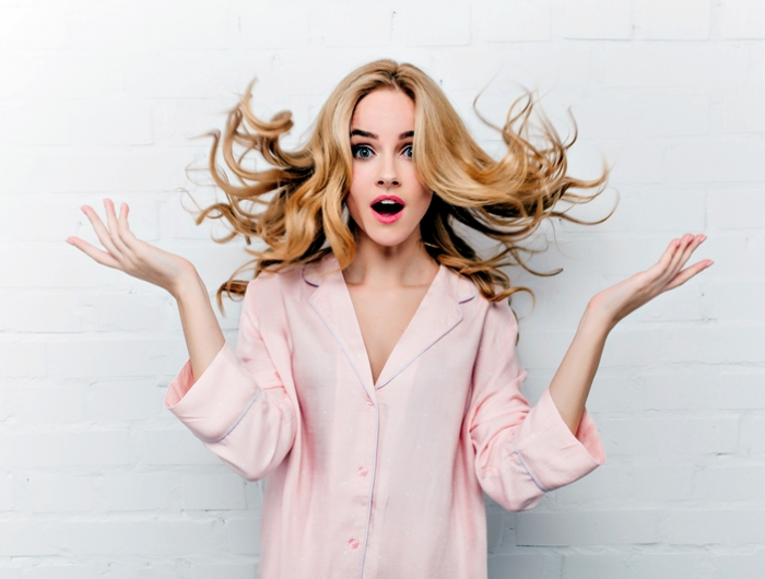 ecstatic blue eyed woman with long blonde hair posing in front of white bricked wall. indoor shot of surprised girl in beautiful pink pyjamas..