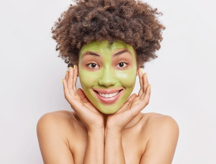 portrait of lovely cheerful young afro american woman keeps hand on face applies green cucumber mask for skin nourishing poses topless against white background. cosmetology and wellbeing concept