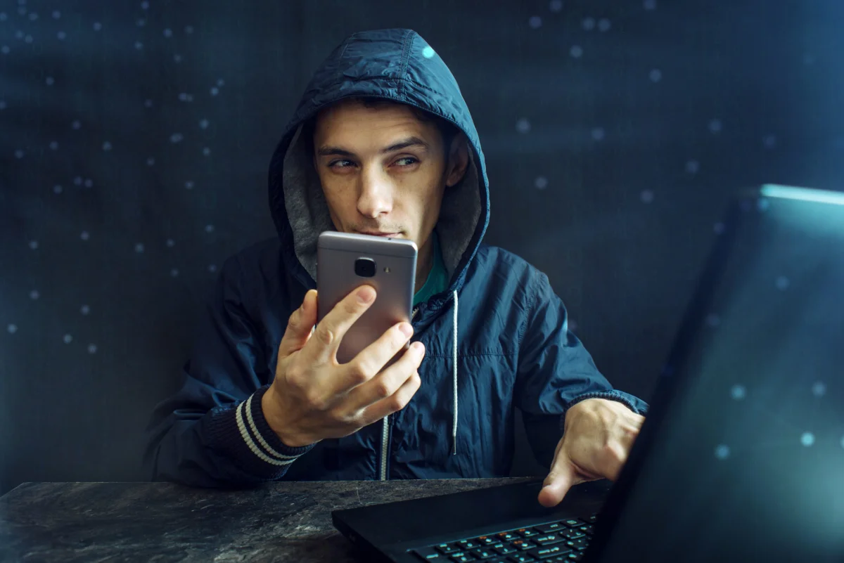 hacker mit blauer jacke hackt smartphone und tippt auf laptop