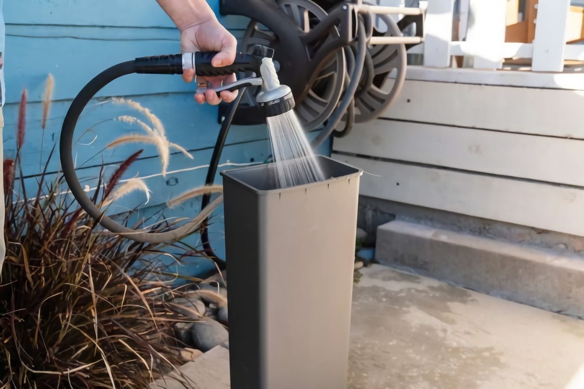 eine frau waescht einen plastikmuelleimer mit einem schwarzen gartenschlauch