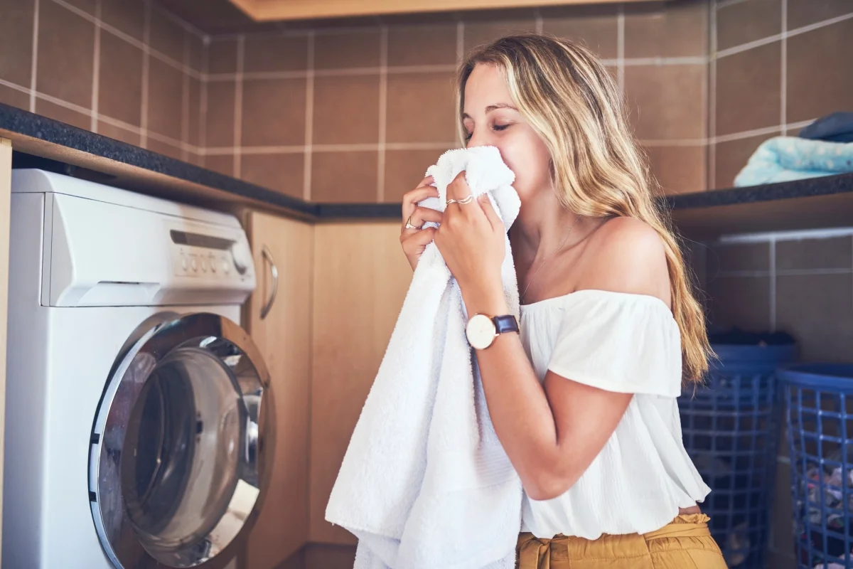 frau mit blondem haar und weißer bluse riecht an frisch gewaschenem handtuch neben der waschmaschine
