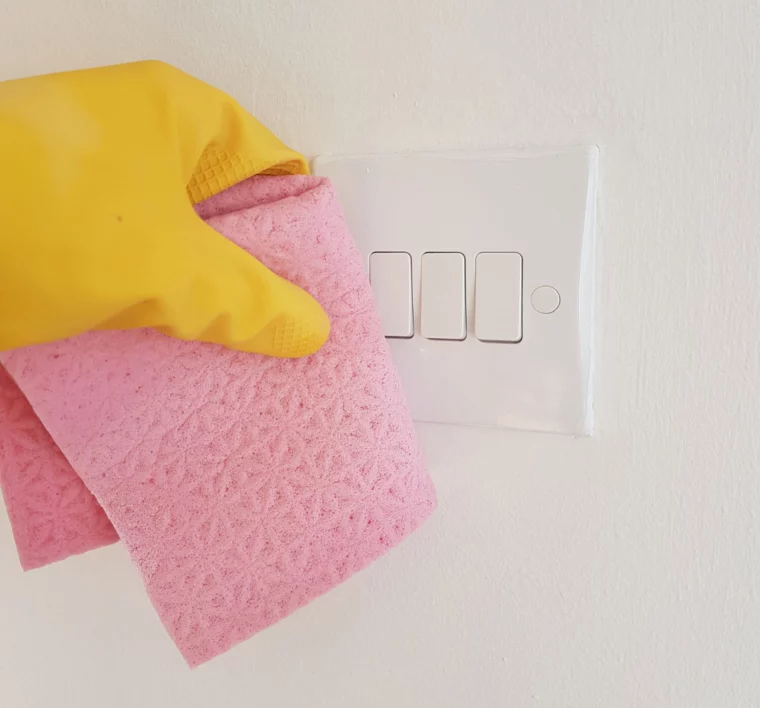frau mit gelbem handschuh reinigt weißen lichtschalter mit rosa tuch an weißer wand