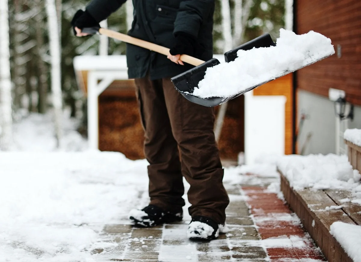 mann mit schwarzer jacke und stiefeln und brauner hose schaufelt schnee vom fliesenboden