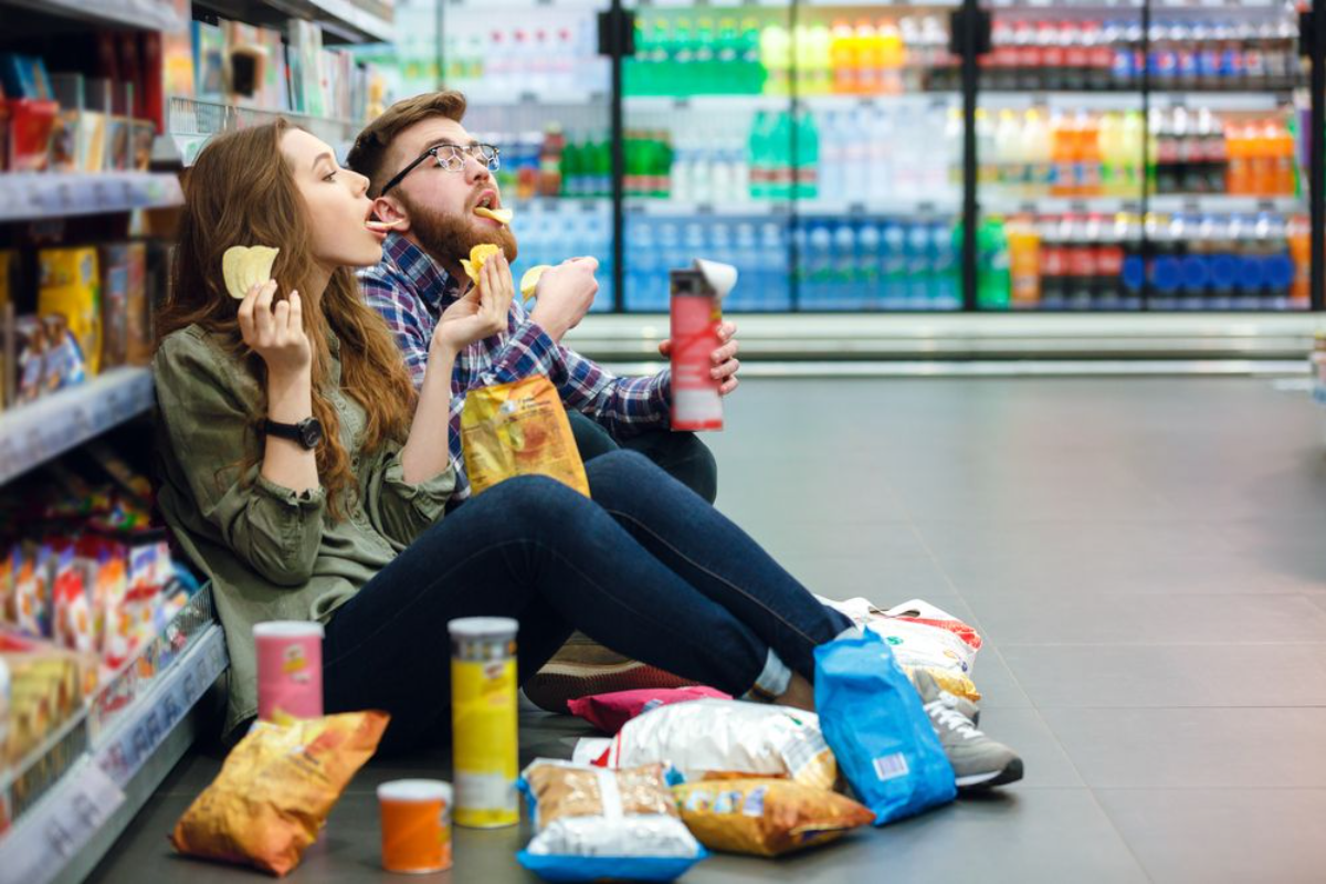 mann und frau sitzen auf dem boden im supermarkt und essen chips
