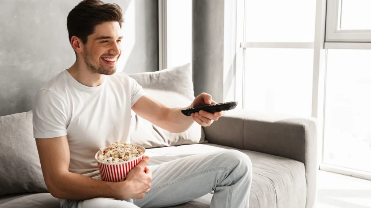was kann man abends naschen ohne zuzunehmen gesunde snacks fuer fernsehabend mann sitzt am sofa laecht sieht fern haelt becher popcorn