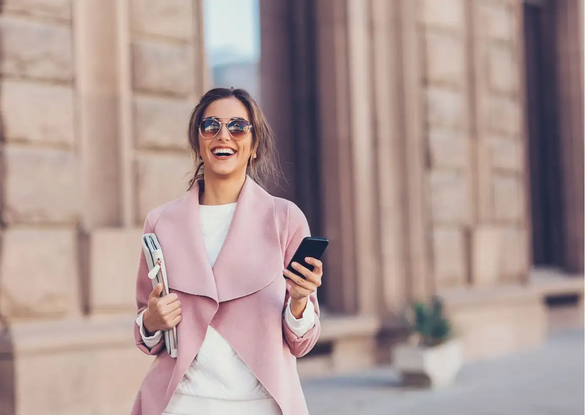 frau mit rosa mantel und weiße kleidung trägt sonnenbrillen trends für 2023 und hält telefon