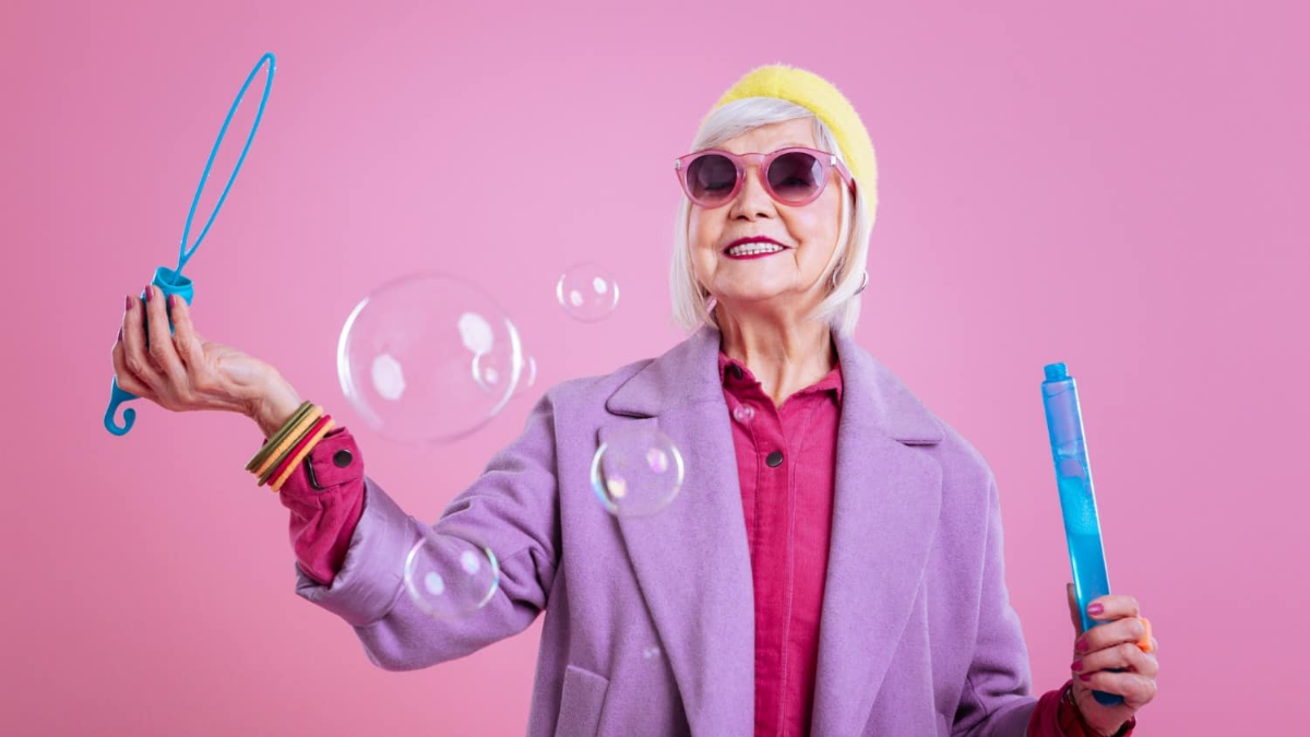 frau nach 60 trägt rosa hemd und lila mantel und rosa sonnenbrille und gelben hut macht seifenblasen auf rosa hintergrund