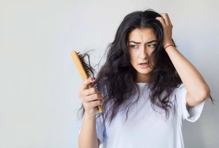 frau mit weißem t shirt und schwarzen haaren hält eine haarbürste und betrachtet die haare die sie verliert