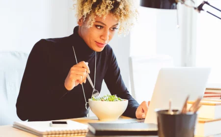 frau mit schwarzem poloshirt und lockigem haar isst salat im büro neben einem laptop