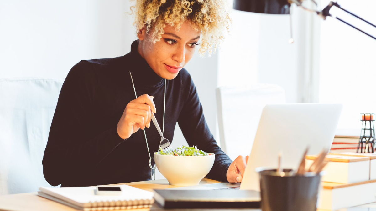 frau mit schwarzem poloshirt und lockigem haar isst salat im büro neben einem laptop