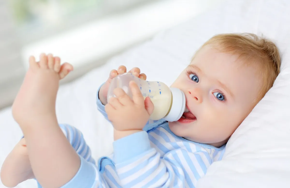 blondes baby mit blauen augen mit gestreiftem strampler trinkt milch babyflasche