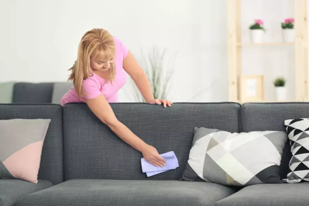 frau mit blondem haar und rosa t shirt reinigt graue couch mit blauem handtuch