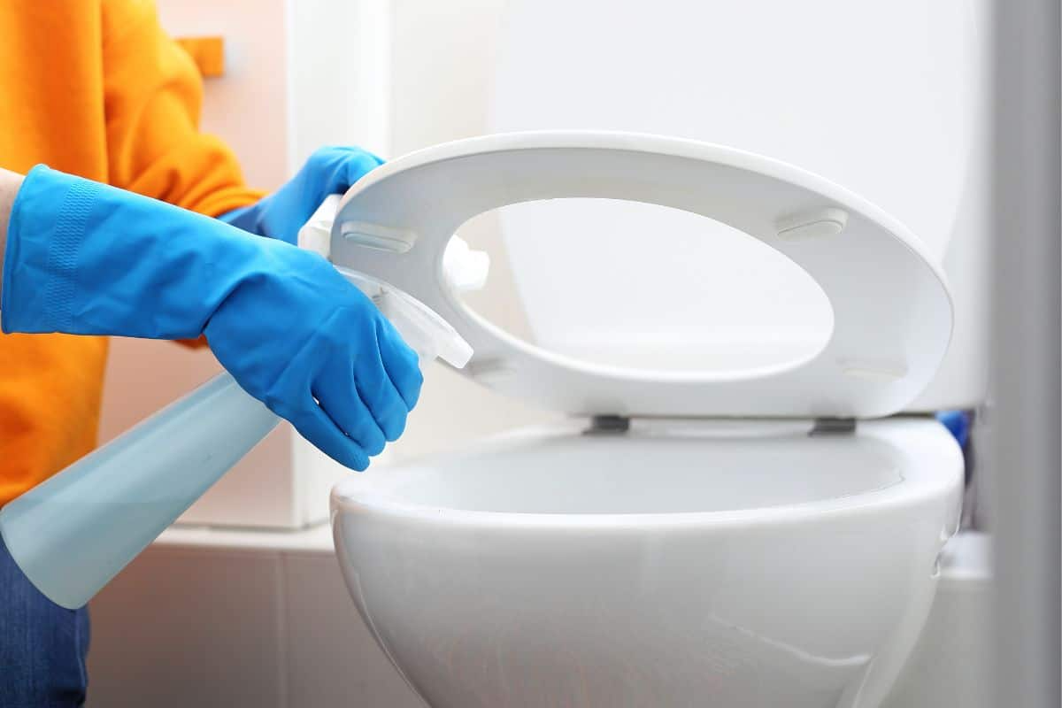 person mit orangefarbener bluse und blauen handschuhen, die die toilette mit desinfektionsmittel besprüht