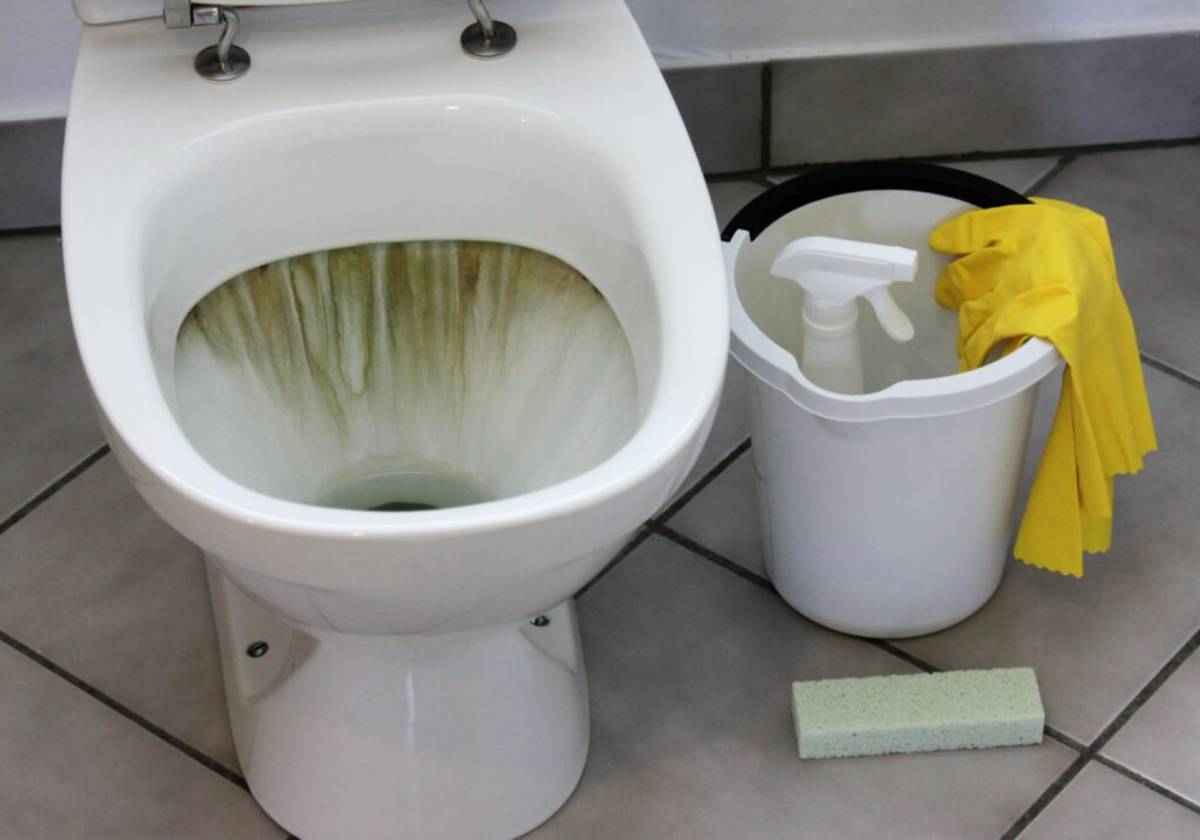 weiße toilette mit urinstein, neben weißem eimer mit gelben handschuhen und schwamm