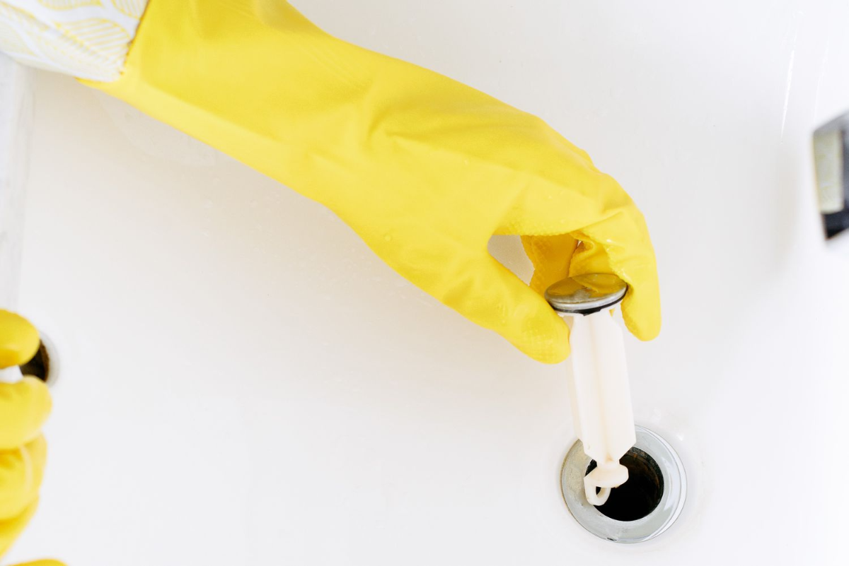 person mit gelben handschuhen, die den abflussstopfen entfernt, um ihn zu reinigen