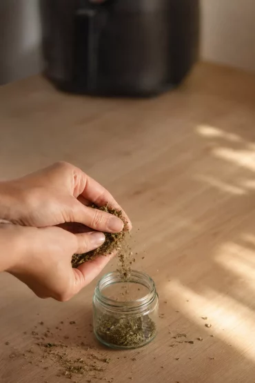 Hände zerbröseln getrocknete Kräuter über einem kleinen Glas